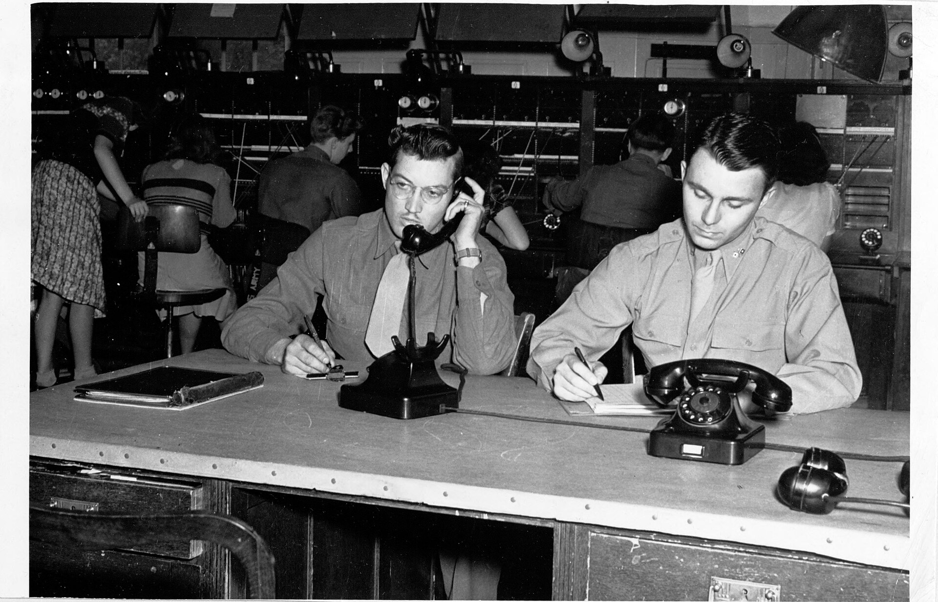 Two operators at a switchboard desk, one taking a call and one writing the details down
