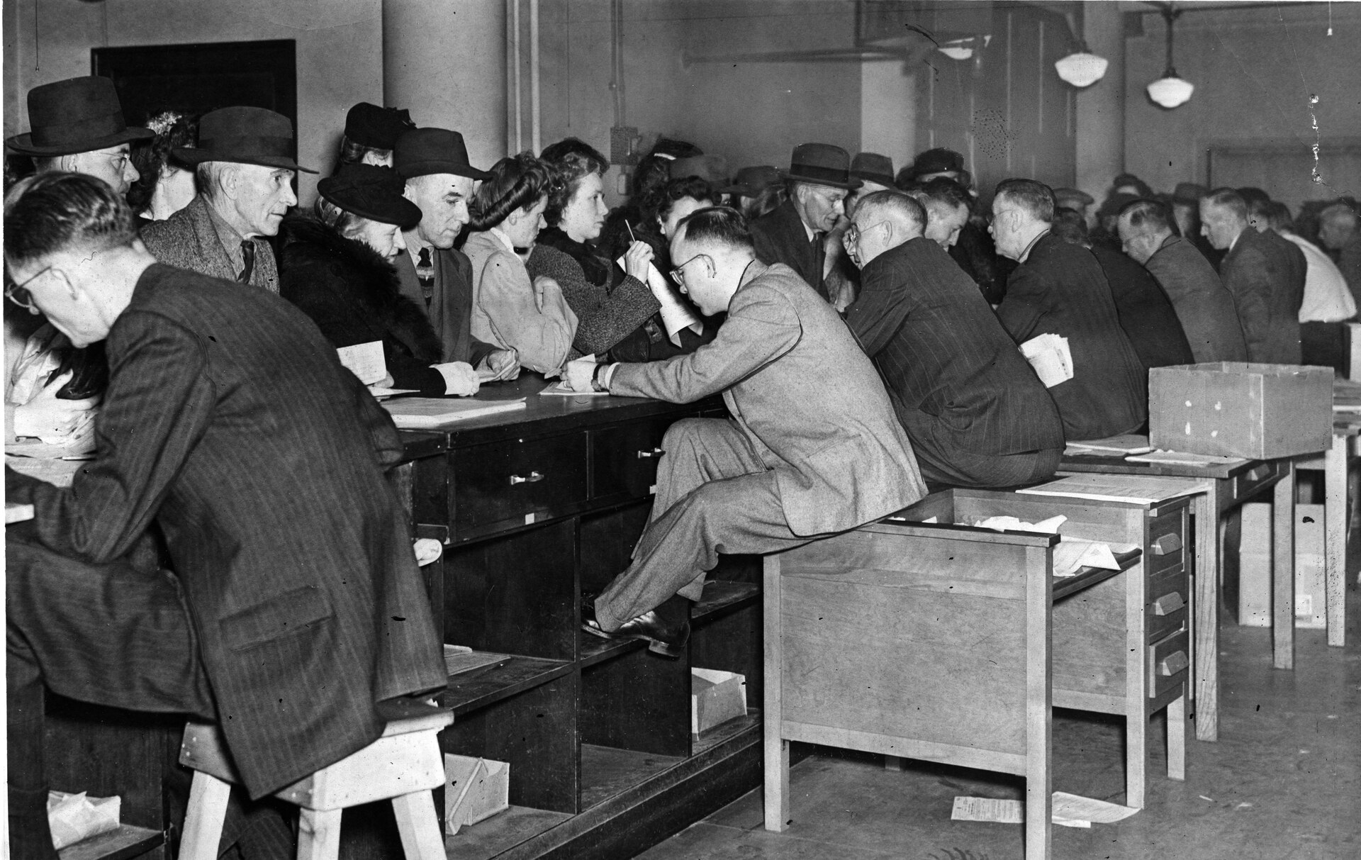Clerks at long desks checking paper forms with customers, 1946