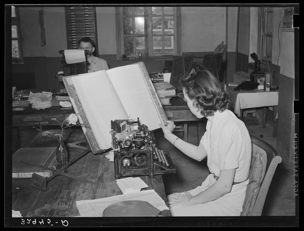 A clerk copying entries from a large ledger on a typewriter in a county office