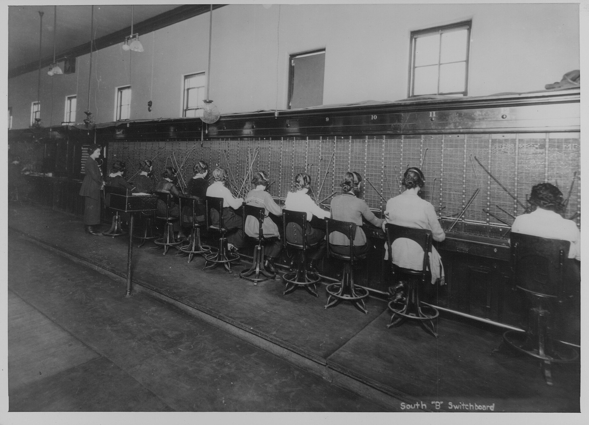 A long telephone switchboard with a row of operators seated at it, early twentieth century